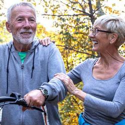 Mann schiebt Fahrrad und geht mit seiner Frau im Wald spazieren, um etwas Gutes für seine Blutzuckerwerte im Alter zu tun.