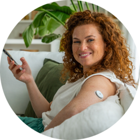 Frau mit CGM-Sensor am Arm hält einen Applikator und hat auf ihrem Smartphone die Accu-Chek SmartGuide App.